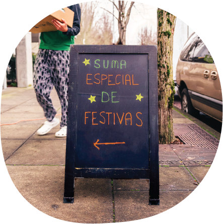 Chalkboard sign reads: suma especial de festivas, an arrow below points to the left. Someone is walking behind the box with a food box